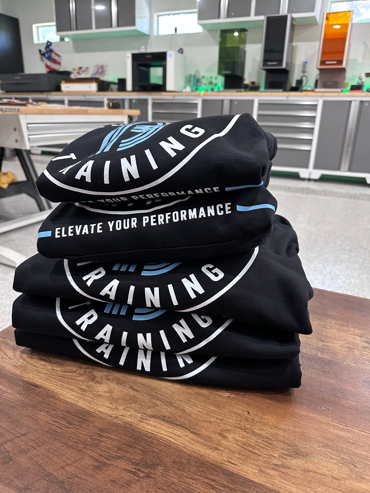 Stack of custom black hoodies featuring white and blue printed training logos, personalized by Cuart Studios in Spring, Texas.